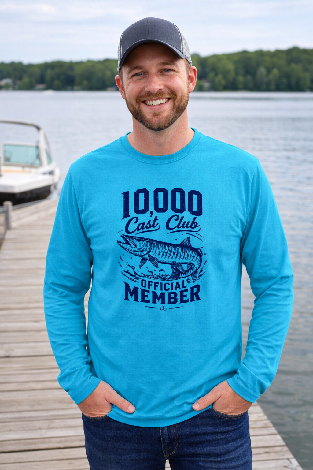 A smiling man wearing a bright turquoise long-sleeve graphic t-shirt and a dark baseball cap stands on a wooden dock with a lake and trees in the background. The shirt features a dark blue graphic that reads '10000 Cast Club OFFICIAL MEMBER' with a fish illustration.